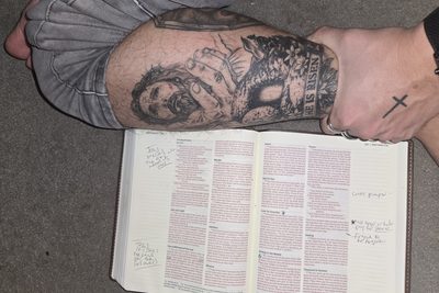 A tattoo of Jesus with the words He is risen, beside an open Bible at Matthew 5 and 6 with handwritten notes in the margins